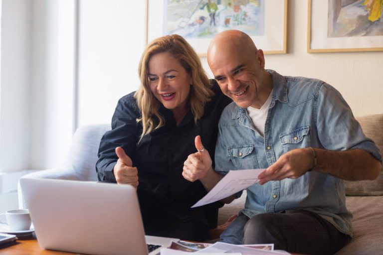 Casal sorrindo em um sofá, fazendo sinal de positivo para um notebook e segurando documentos sobre as janelas de competência do empréstimo CLT da 321bank